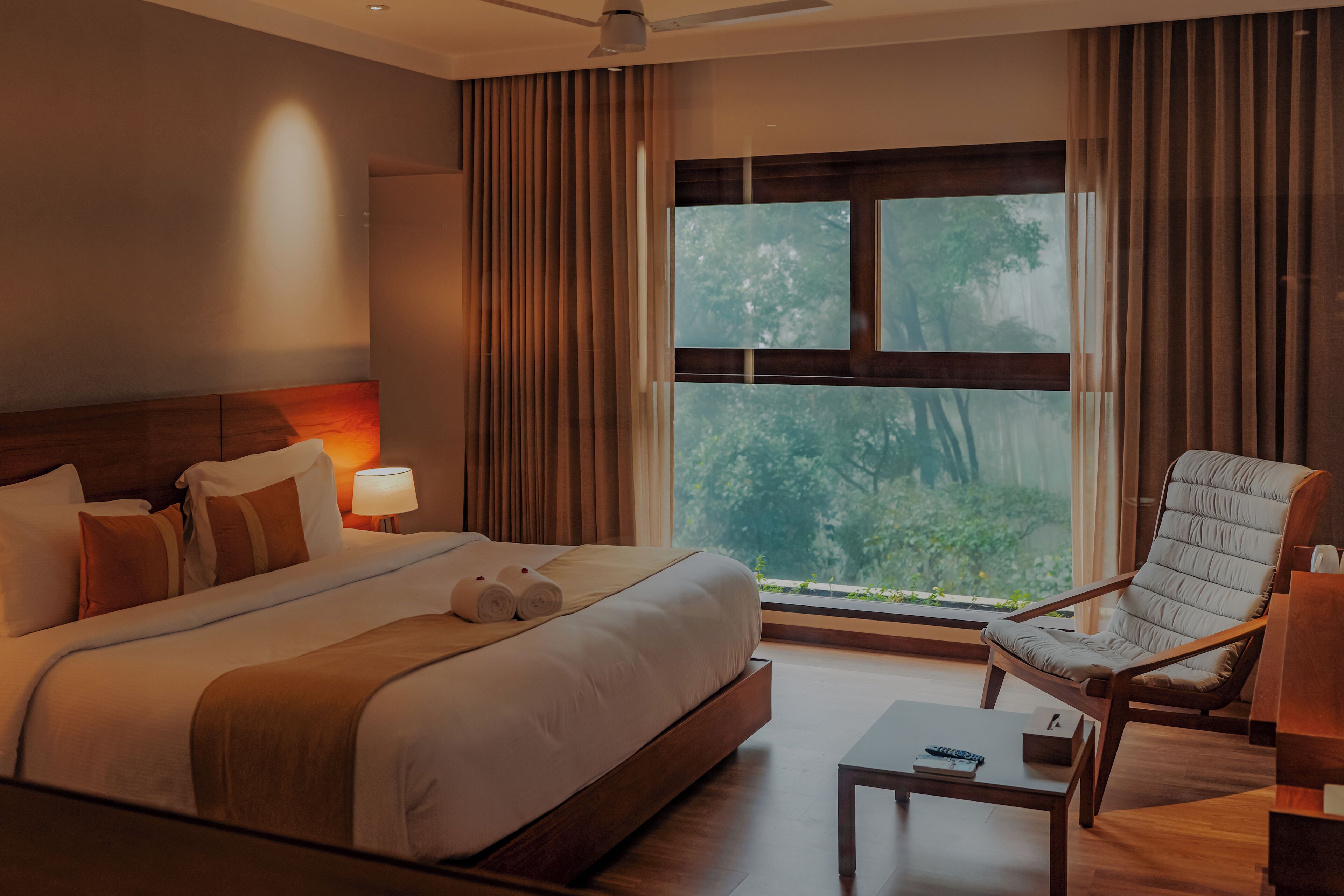 A hotel room with a large bed, a sitting area with a chair, a table, and a view of trees through the window.