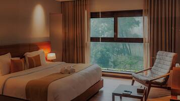 A hotel room with a large bed, a sitting area with a chair, a table, and a view of trees through the window.