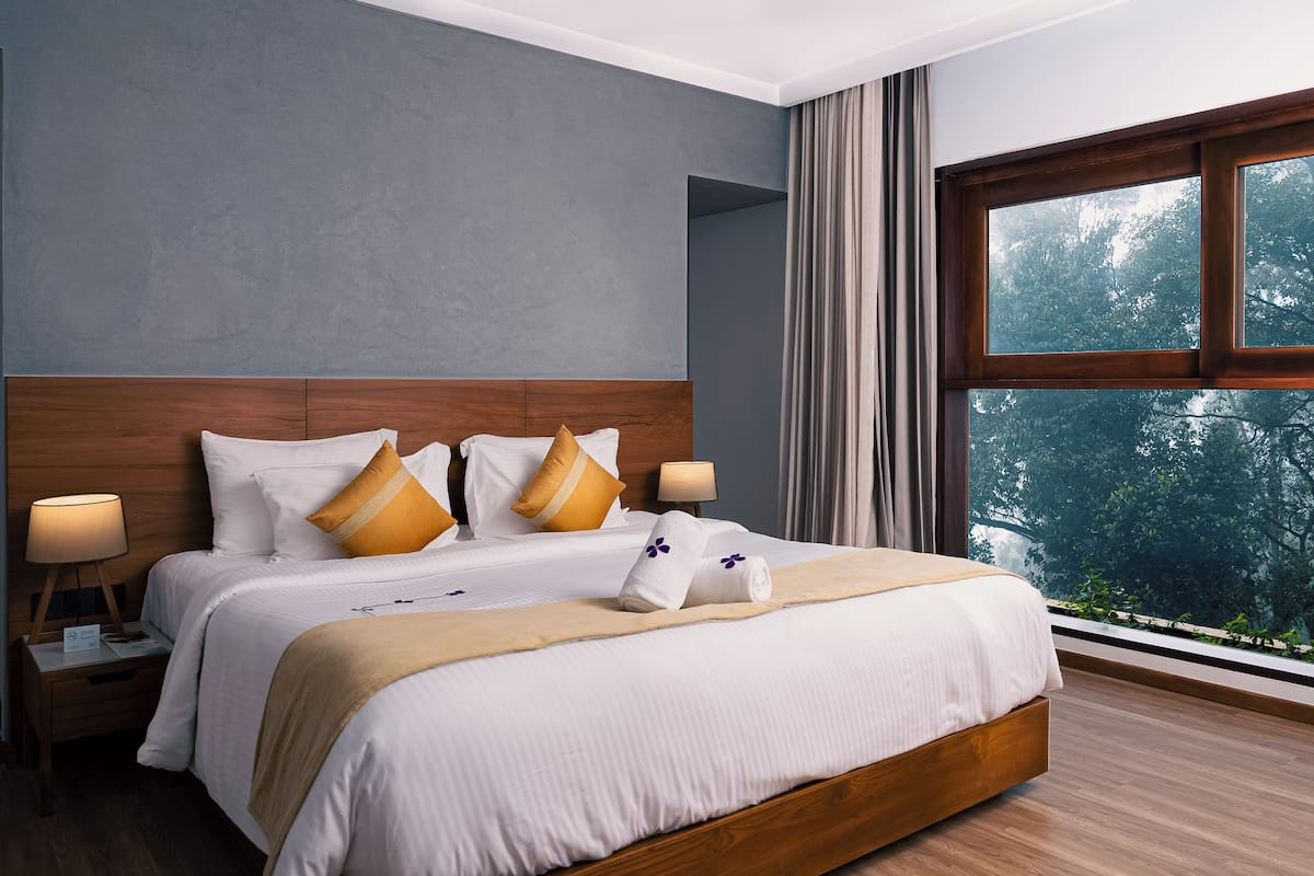 A neatly made bed with white linens and two decorative pillows, a wooden headboard, a bedside table with a lamp, and a large window with a view of trees.