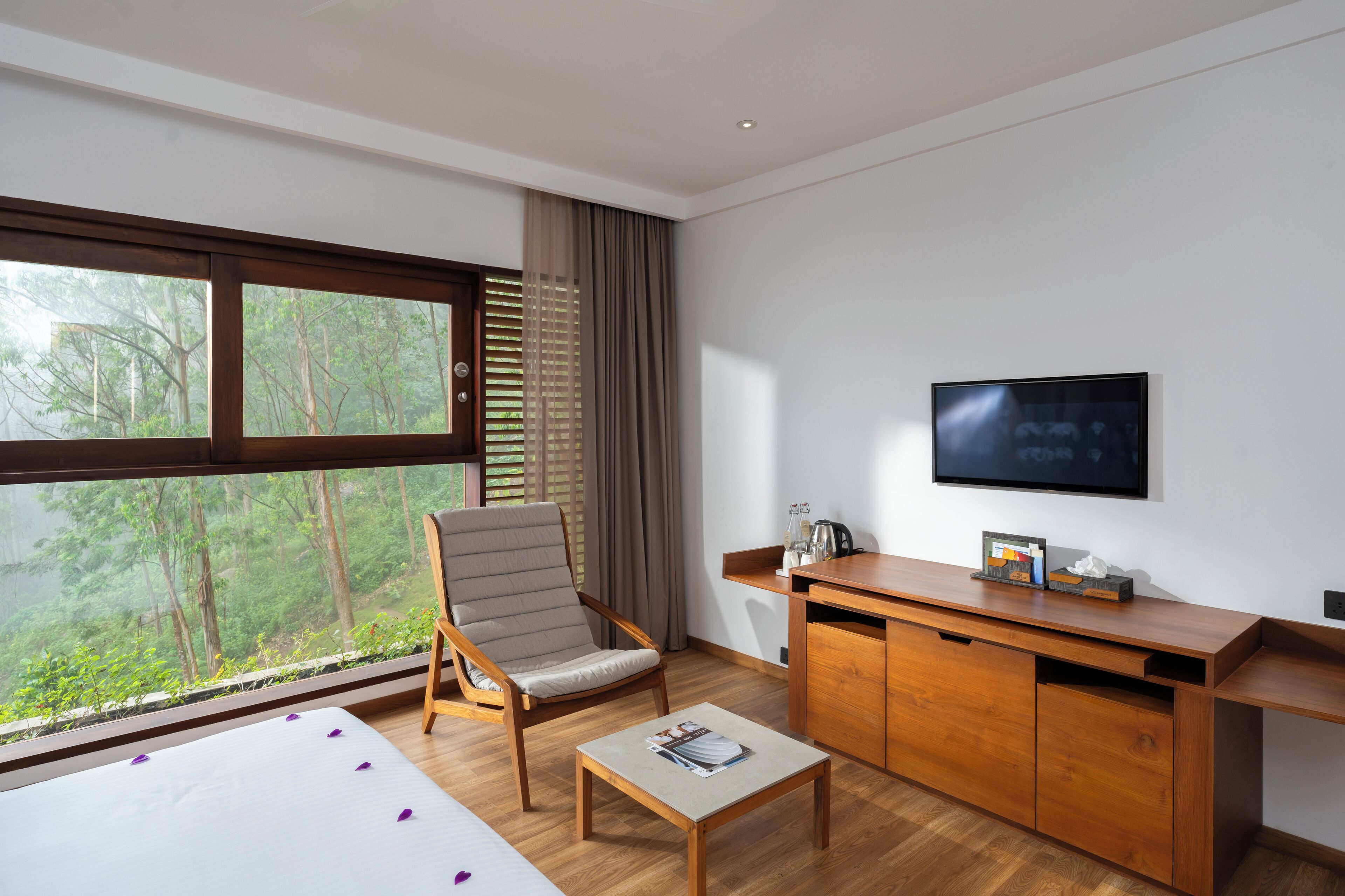 A modern hotel room with a large window, a wooden desk, a television, and a comfortable chair.