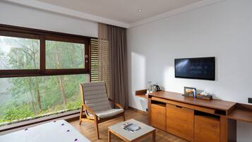 A modern hotel room with a large window, a wooden desk, a television, and a comfortable chair.