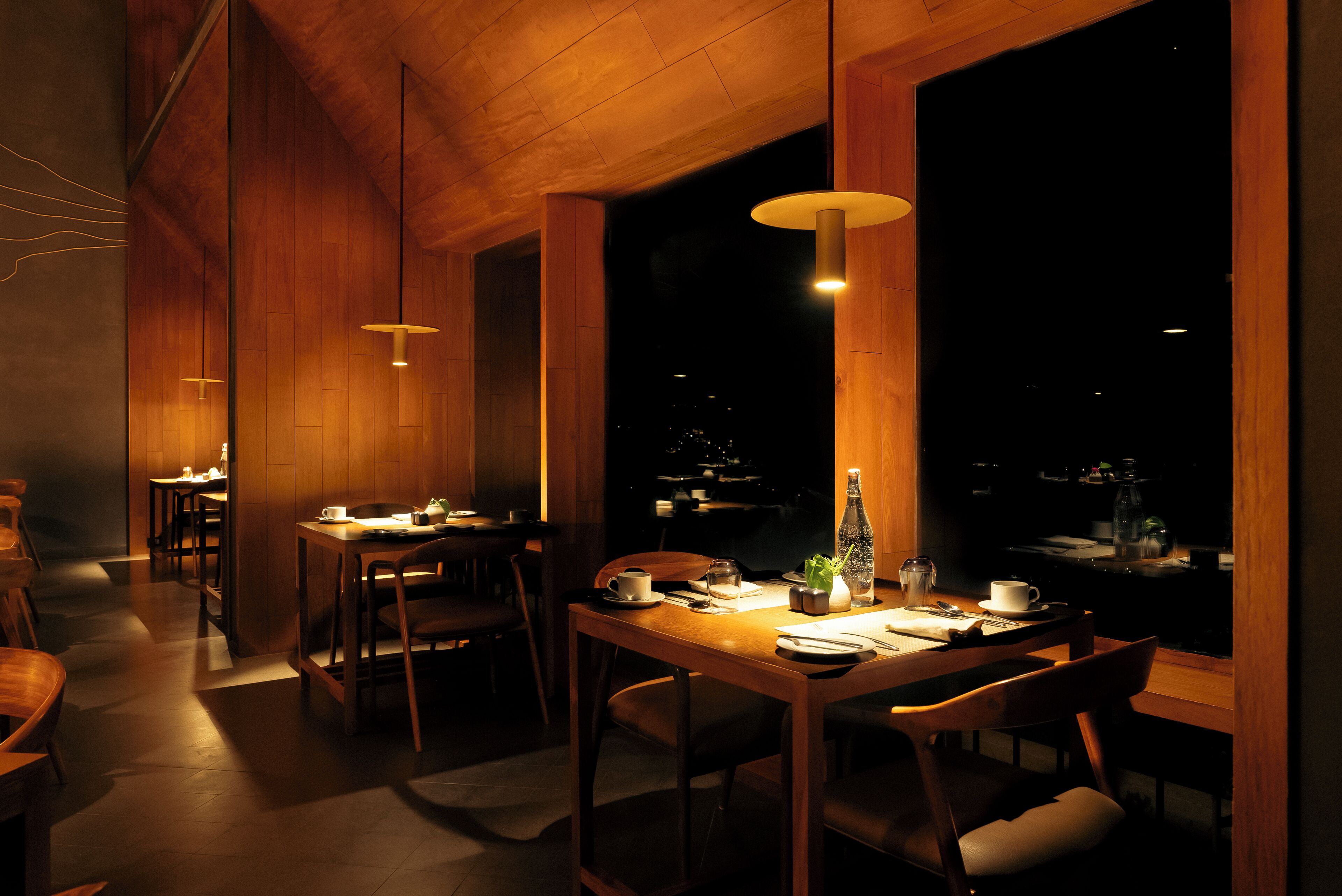 Un restaurante tenuemente iluminado con mesas y sillas de madera, lámparas colgantes y vistas de la ciudad por la noche.