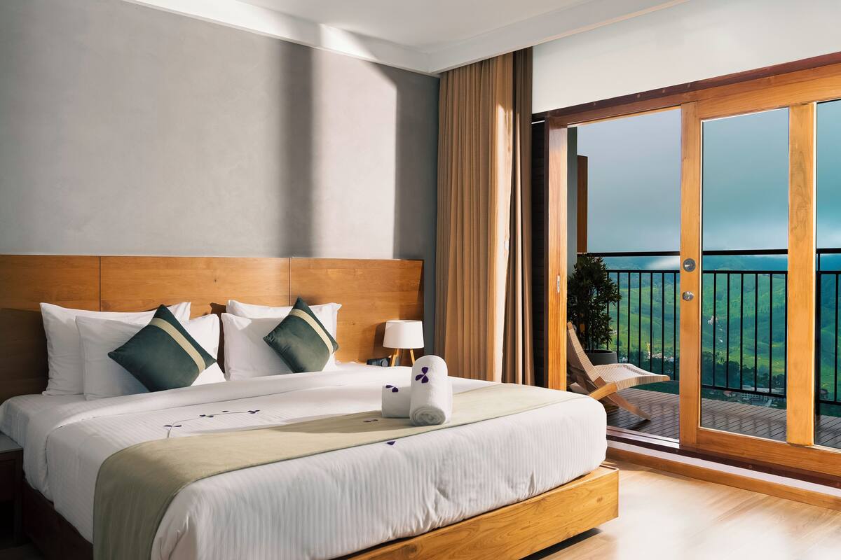 A hotel room with a large bed, a wooden headboard, a balcony with a view, and a sliding glass door.