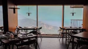 A restaurant with tables set for dining, large windows offering a mountain view, and pendant lighting. - The Valle by Kondody Hotels (Devikolam)