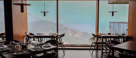 Un restaurante con mesas dispuestas para comer, amplias ventanas que ofrecen vistas a la montaña e iluminación colgante.