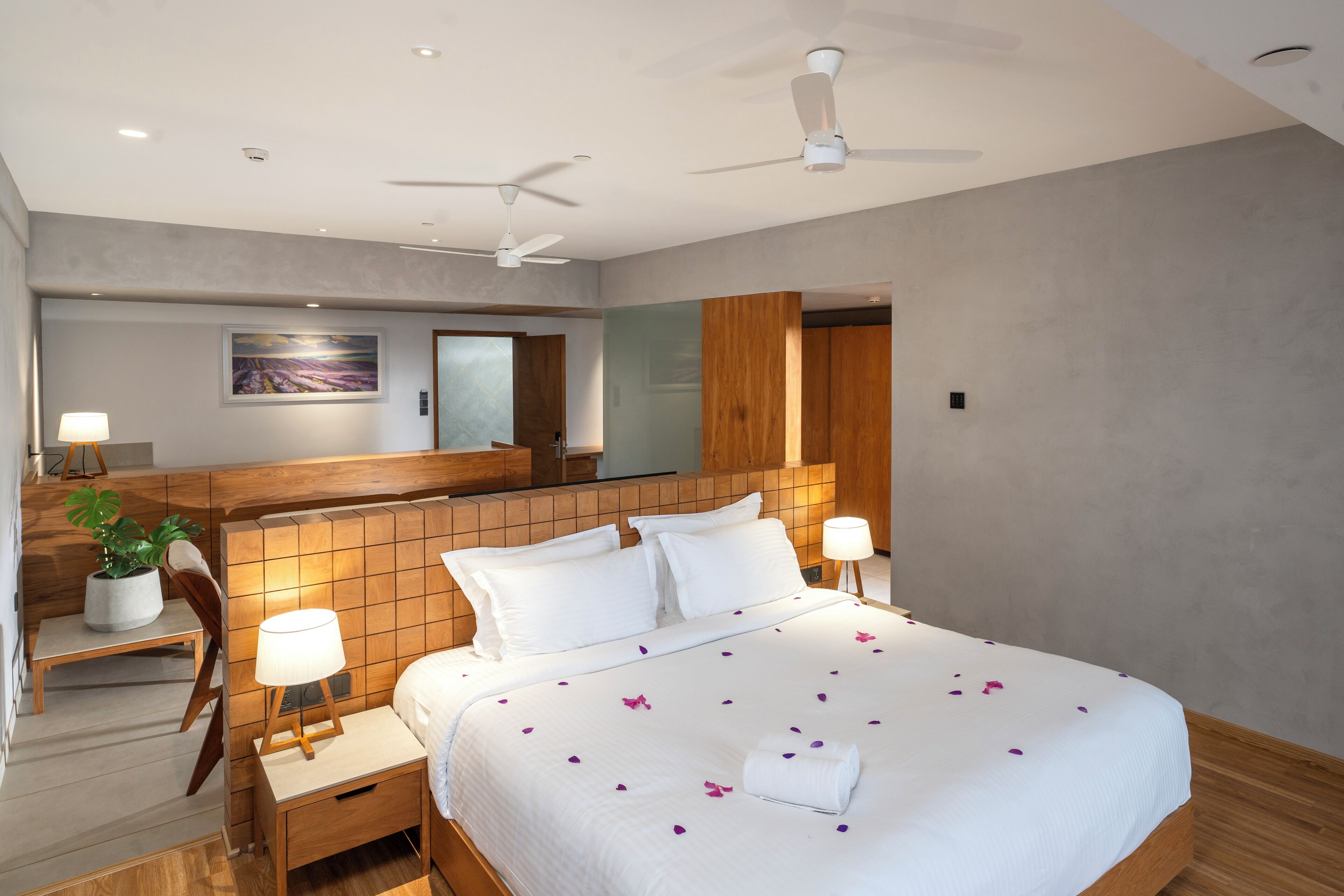A hotel room with a large bed, two bedside tables with lamps, a ceiling fan, a wooden headboard, a potted plant, and a painting on the wall.