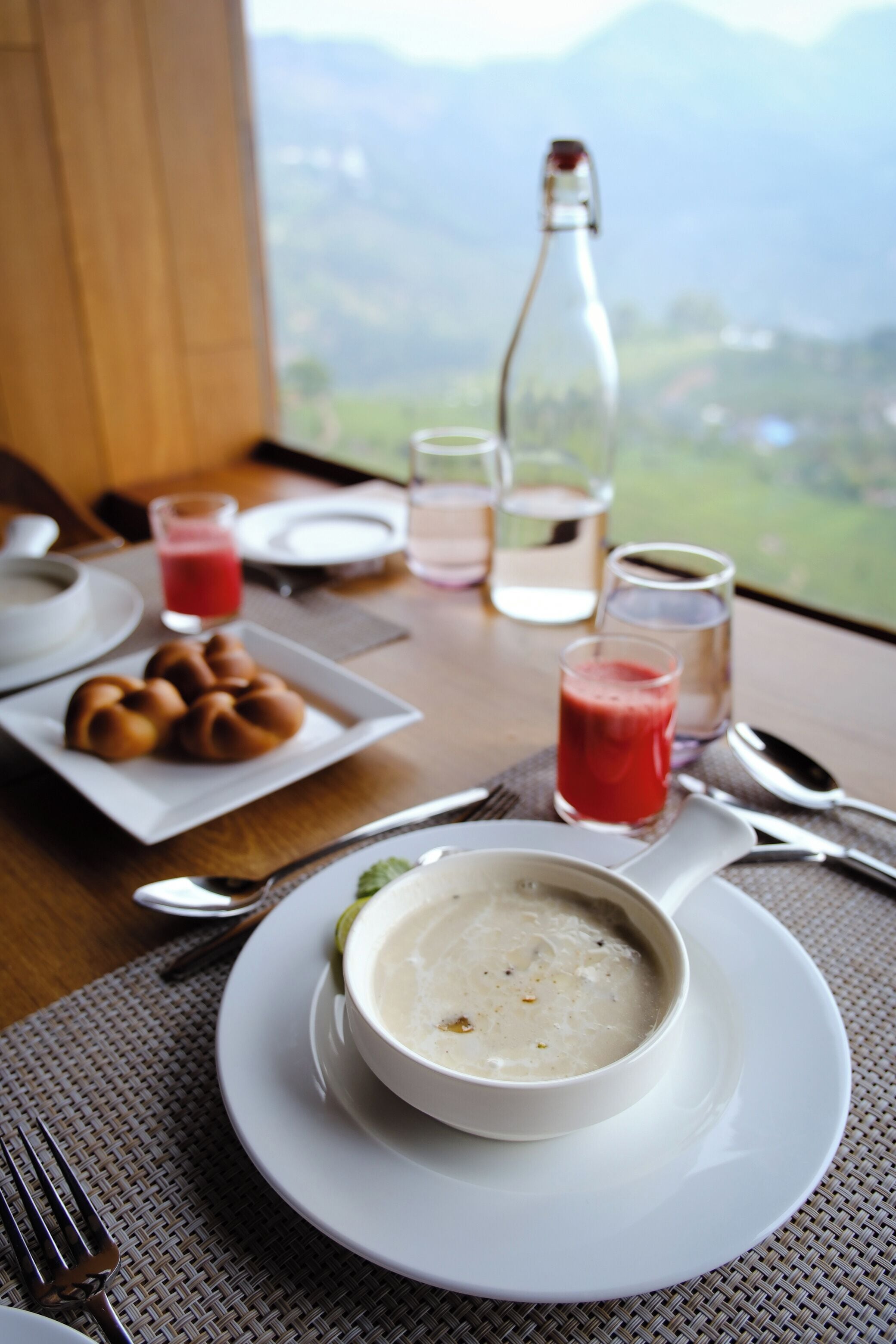 Une table dressée pour le petit-déjeuner, avec un bol de soupe, des croissants et une bouteille de jus, le tout placé devant une vue sur les montagnes.