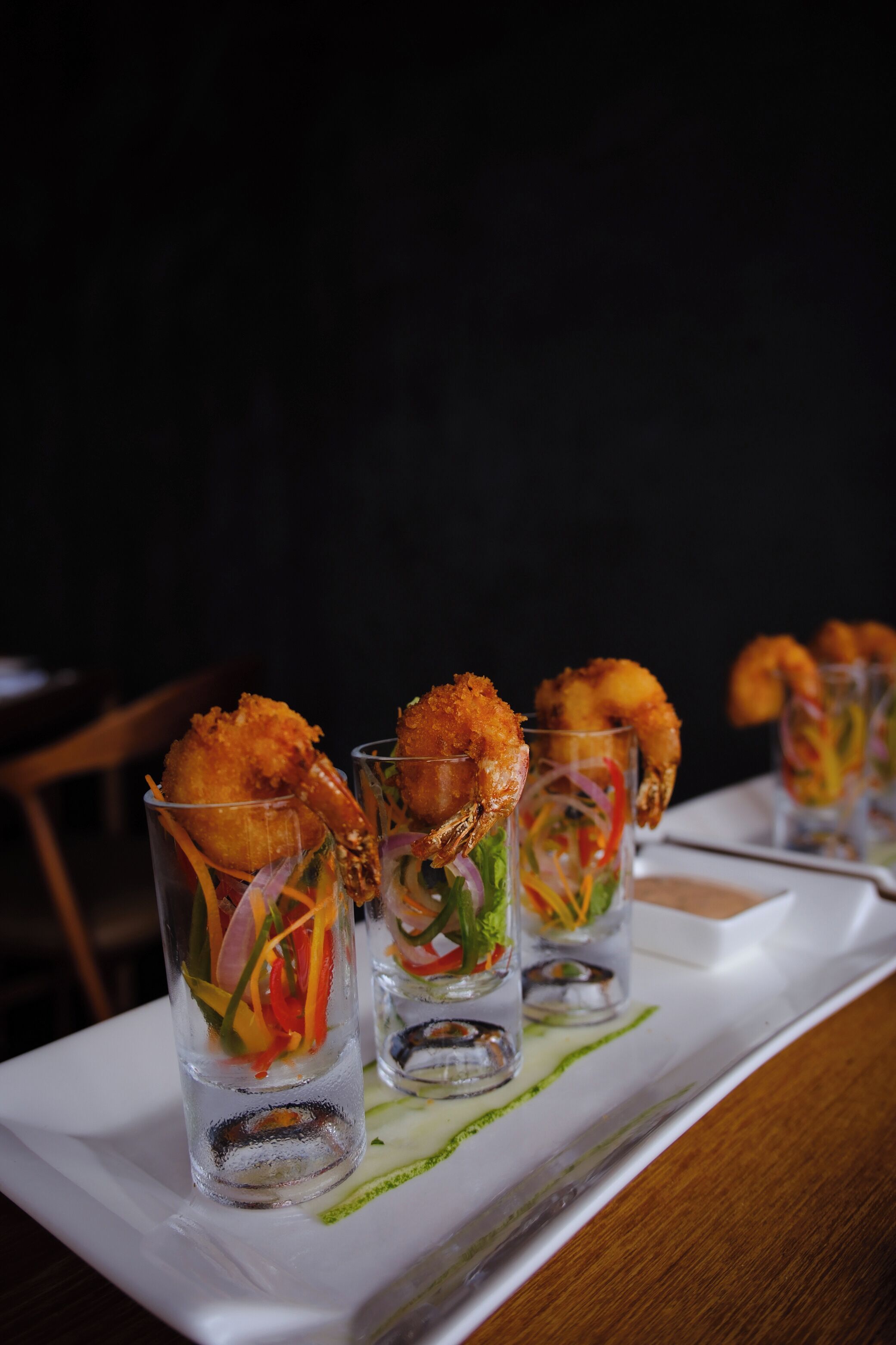 Una hilera de vasos transparentes con aperitivos de camarones y verduras sobre una bandeja blanca.