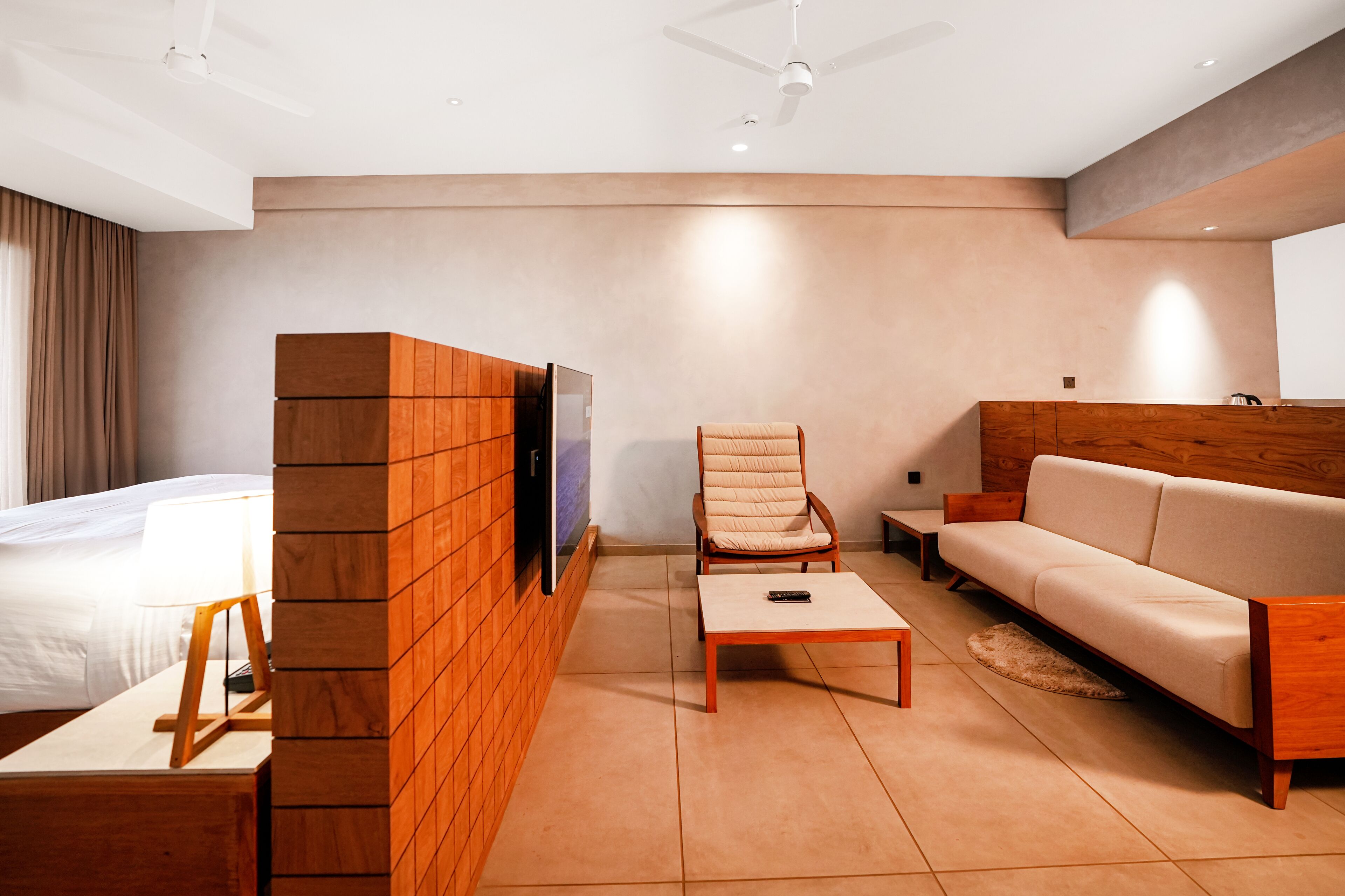 A modern hotel room with a bed, a wooden entertainment unit, a sofa, a chair, and a coffee table.