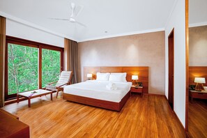 A spacious hotel room with a large bed, wooden flooring, a sitting area with a chair and table, and a view of greenery through large windows. - The Valle by Kondody Hotels (Devikolam)