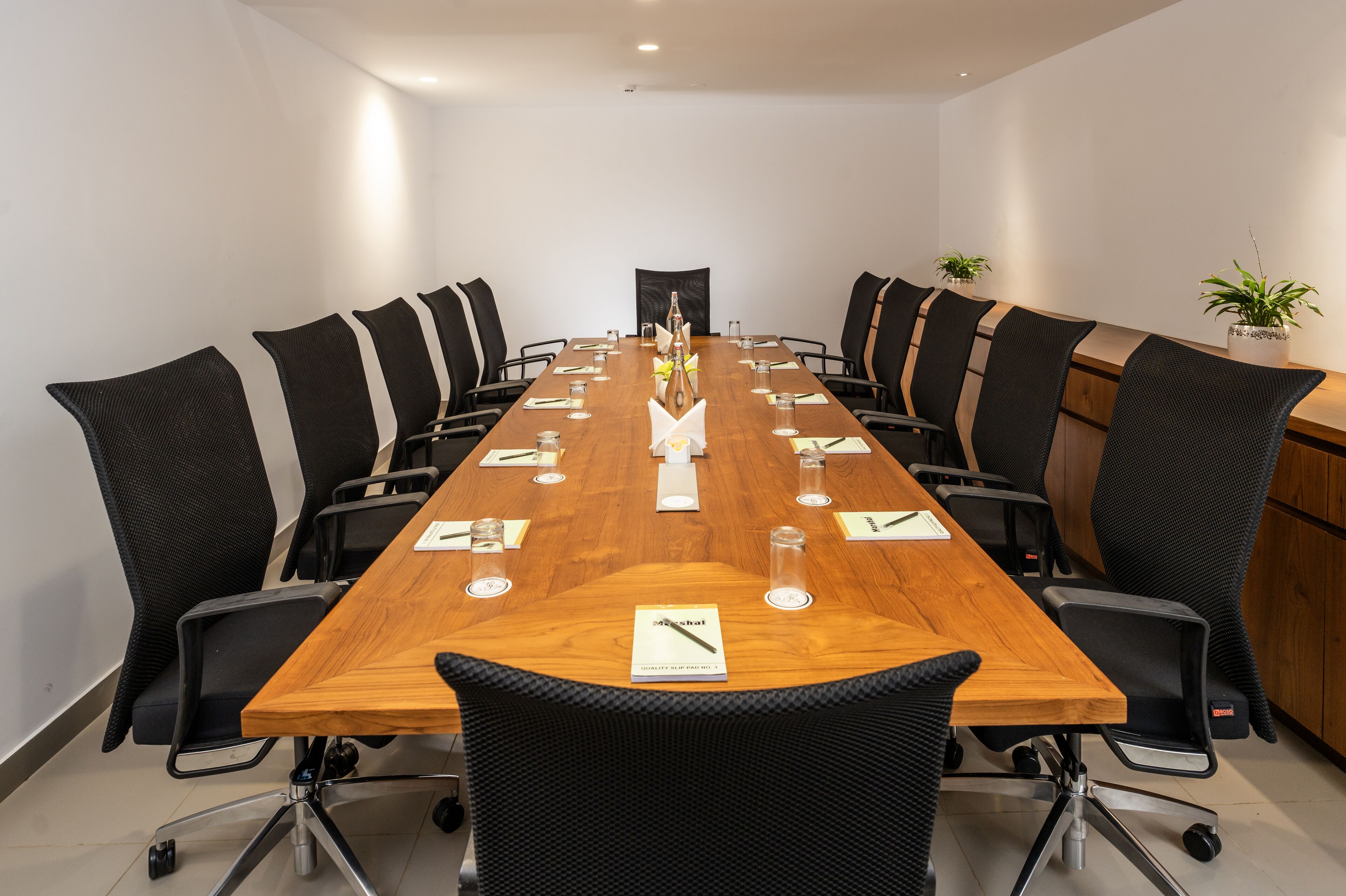 A conference room with a long wooden table, black chairs, and potted plants.