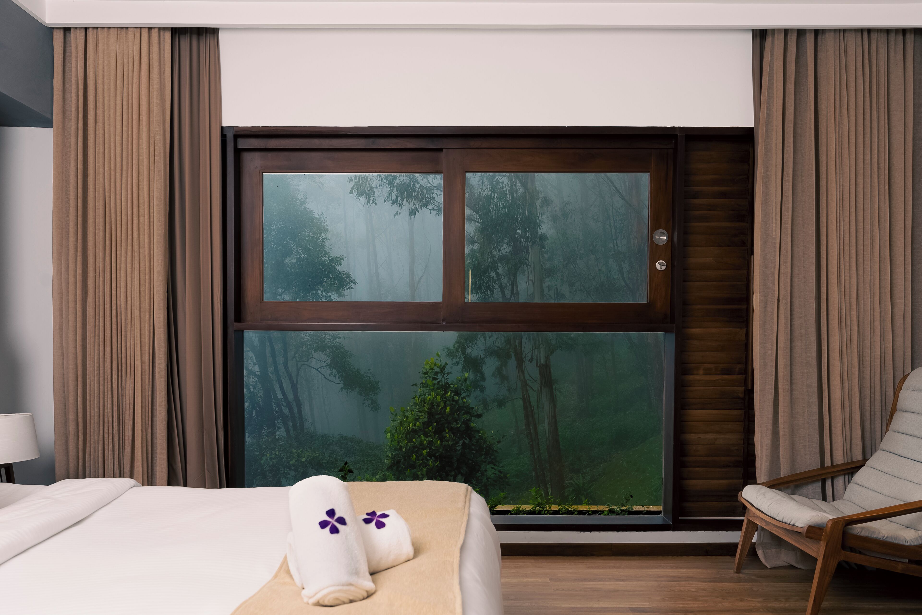 A hotel room with a large window, a bed with towels, a chair, and a nightstand with a lamp.