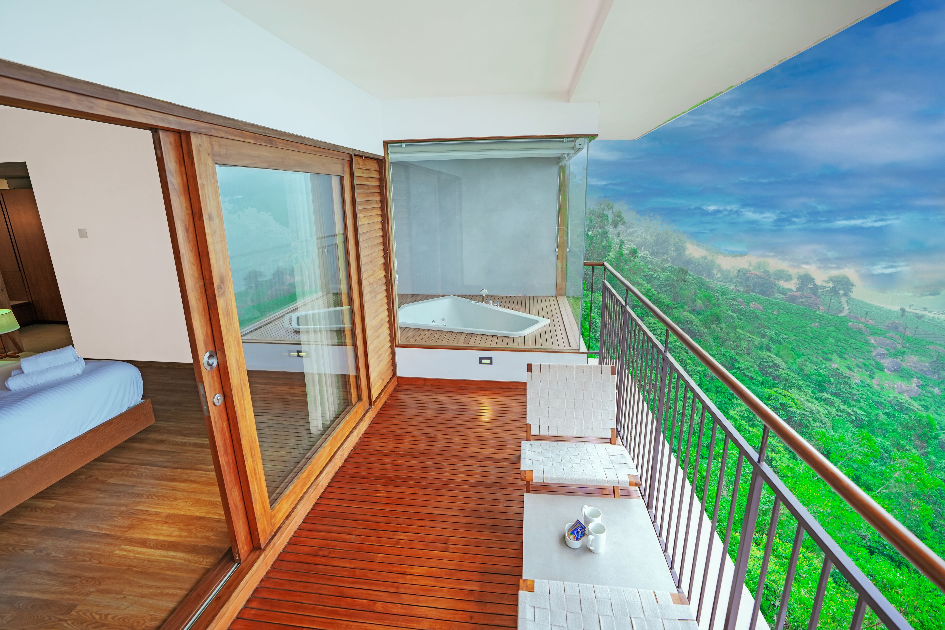 A balcony with a wooden floor, a glass door, a white sofa, a small table with cups, and a scenic view of a green landscape.