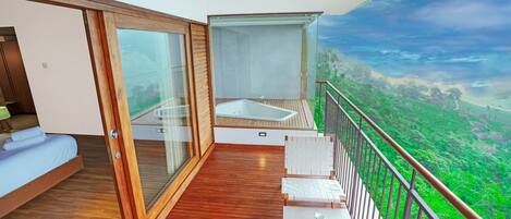 A balcony with a wooden floor, a glass door, a white sofa, a small table with cups, and a scenic view of a green landscape.