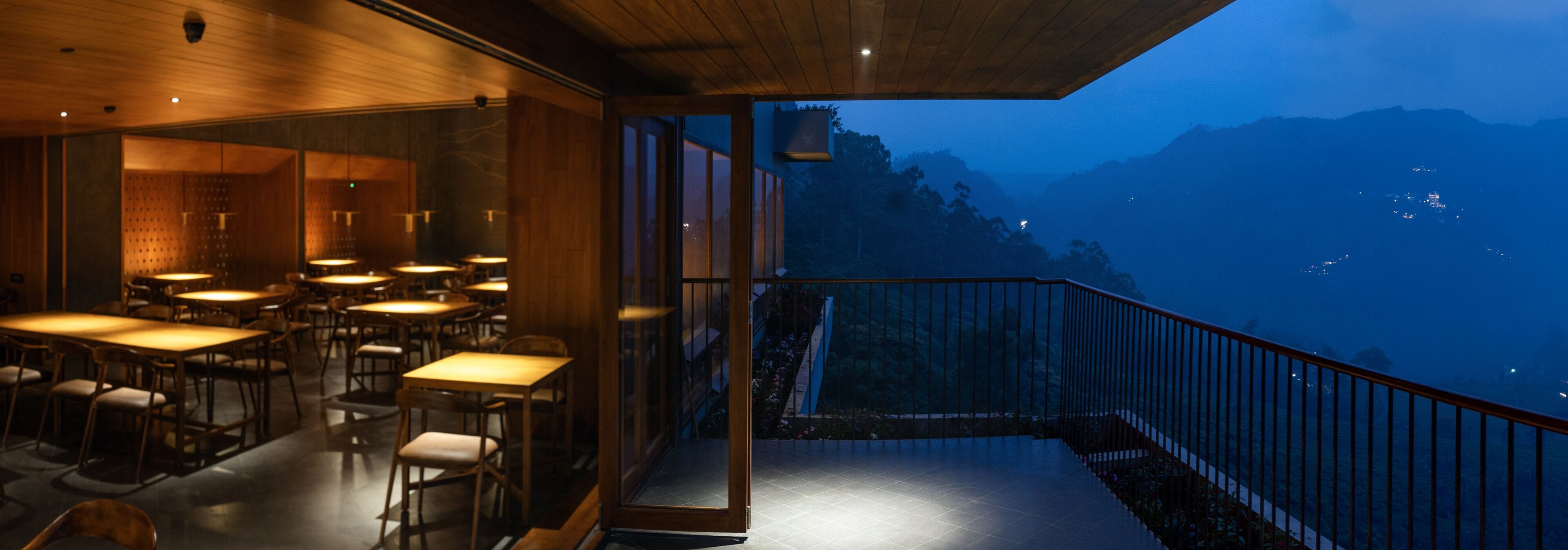 A restaurant with tables and chairs, a balcony with a railing, and a view of mountains in the background.