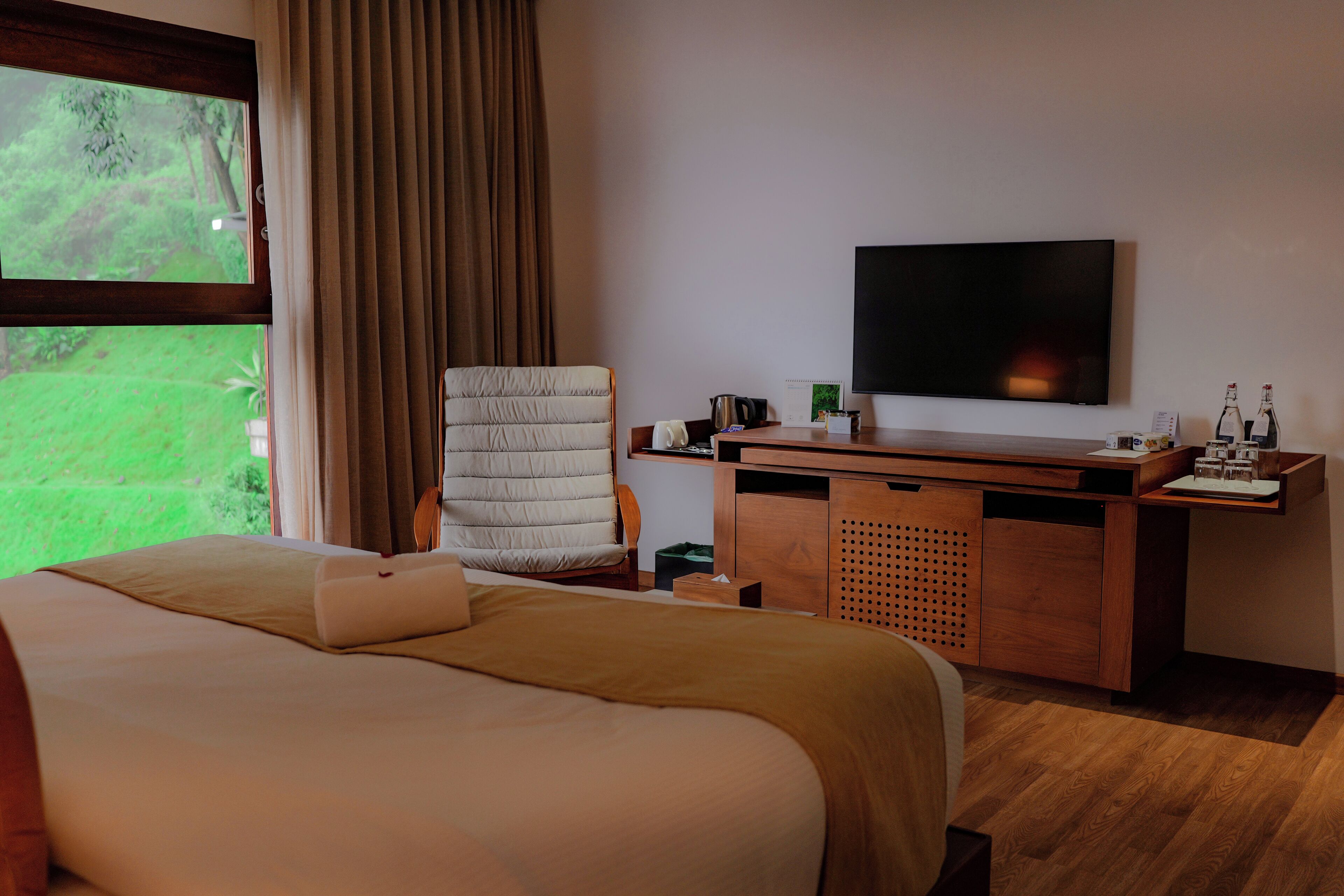 Habitación de hotel con cama, escritorio, silla, televisión y ventana con vista a follaje.