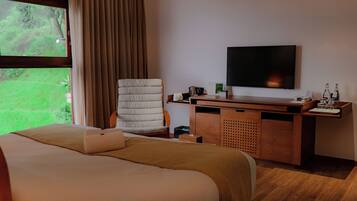 Habitación de hotel con cama, escritorio, silla, televisión y ventana con vista a follaje.