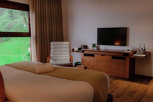Habitación de hotel con cama, escritorio, silla, televisión y ventana con vista a follaje.