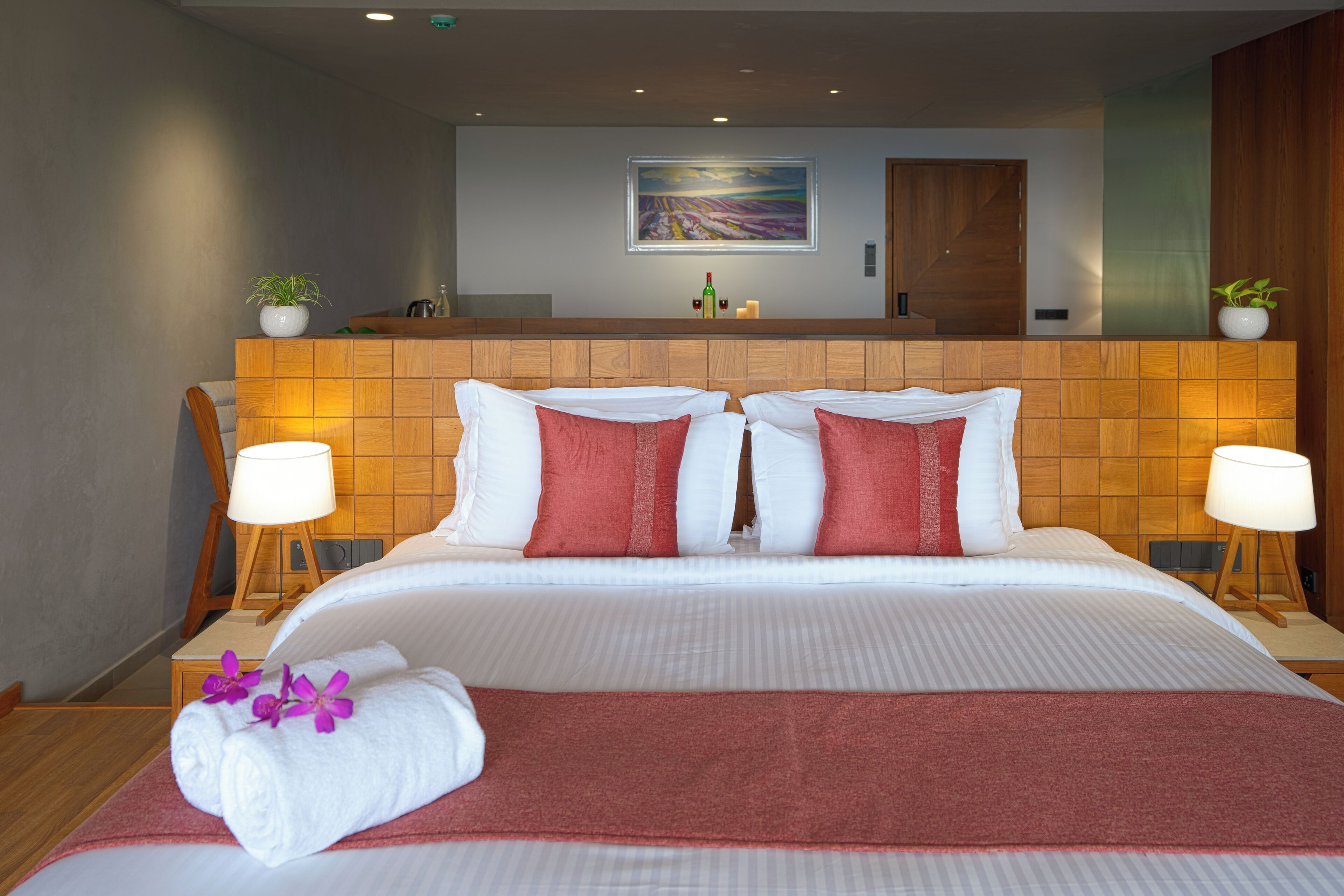 A hotel room with a large bed, two bedside tables with lamps, a wooden headboard, a painting on the wall, and a small plant on the headboard.