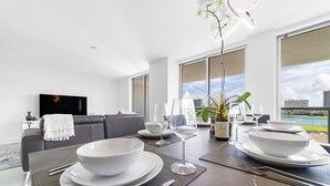 Ein moderner Essbereich mit einem großen Tisch, der für eine Mahlzeit gedeckt ist, einer Couch im Hintergrund und einem Blick auf die Stadt durch große Fenster.