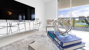 Ein modernes Wohnzimmer mit großem Fernseher, einem weißen Sofa, einem Glastisch und Blick ins Freie.