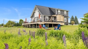 A modern two-story house with a covered deck, surrounded by a lush garden and purple flowers. - Heron House, Beautiful seaside retreat close to Cavendish (North Rustico)