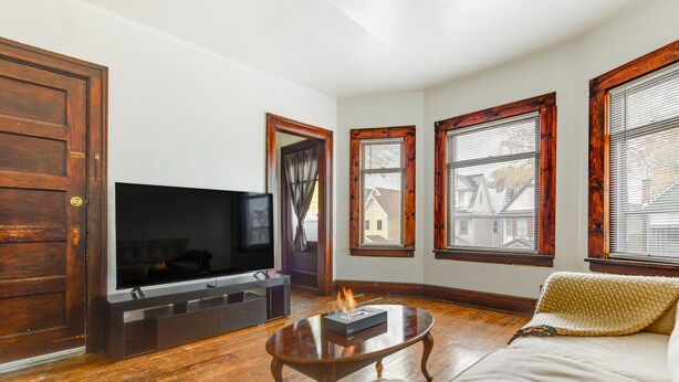 A living room with a flat-screen TV, wooden flooring, a fireplace, and large windows with shutters. - Charming Buffalo Apartment: 2 Mi to Downtown (Buffalo)