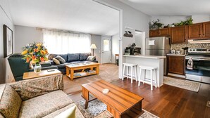 Uma sala ampla com sofá, mesinha de centro e um espaço de jantar com banquetas. Cozinha com armários de madeira e eletrodomésticos visíveis.