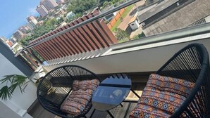 A balcony with two black wicker chairs and a small table, overlooking a cityscape with buildings and greenery. - Beautiful & Spacious 1 BR Suite - Modern African Decor & Ocean View, Osu (Accra)