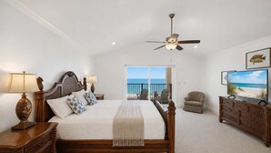 A bedroom with a bed, a ceiling fan, a television, and a view of the ocean through a balcony door.