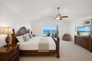 Quarto com cama, ventilador de teto, televisão e vista para o oceano através de uma porta de varanda.