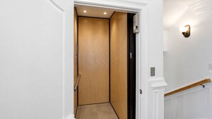 A hallway with a wooden door leading to an elevator, white walls, and a light fixture on the wall.
