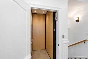 Corredor com porta de madeira que dá para um elevador, paredes brancas e luminária na parede.