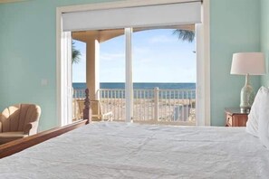 A bedroom with a large bed, a chair, a nightstand, and a view of the beach through a sliding door.