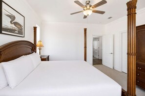 Quarto de hotel com uma cama grande, um ventilador de teto, cabeceira de madeira, mesa de cabeceira com luminária e um quadro na parede.