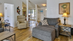 Un salon avec un canapé gris, un fauteuil beige et une table basse en bois. On aperçoit une lampe sur une table d’appoint et un tableau encadré au mur.