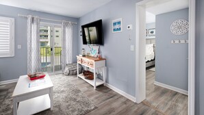 A modern bedroom with a TV, a wooden dresser, a large window with curtains, and a view of a balcony.