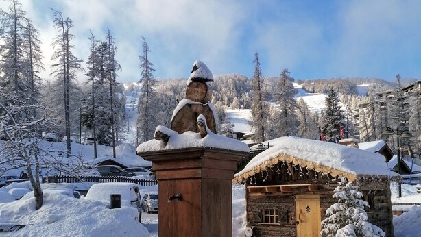 Ein Steinkunstbrunnen mit Statue, eine Holzhütte und schneebedeckte Bäume in einer Berglandschaft.