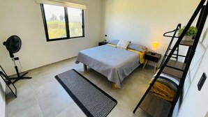 A bedroom with a bed, a standing fan, a bedside table with a lamp, a window, and a black ladder. - La Ventana Apartments Depa #4 (La Ventana)