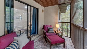 Ein Balkon mit Holzboden, ein gepolsterter Stuhl, ein kleiner Tisch mit einer Lampe und Blick auf Bäume durch eine Glastür.