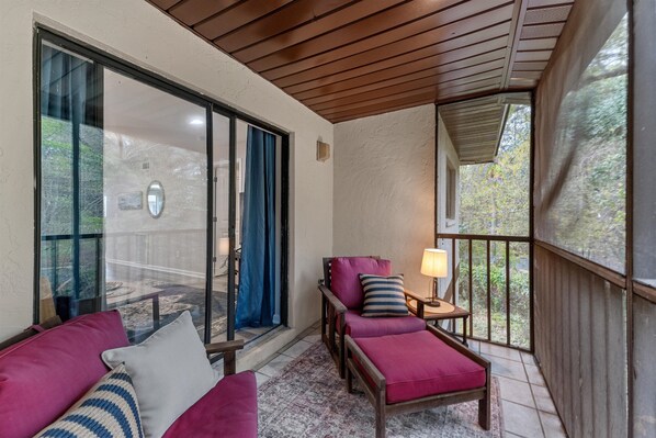 Un balcon doté d’un plafond en bois, d’un fauteuil rembourré, d’une petite table avec une lampe et d’une vue sur les arbres, visible à travers une porte vitrée.
