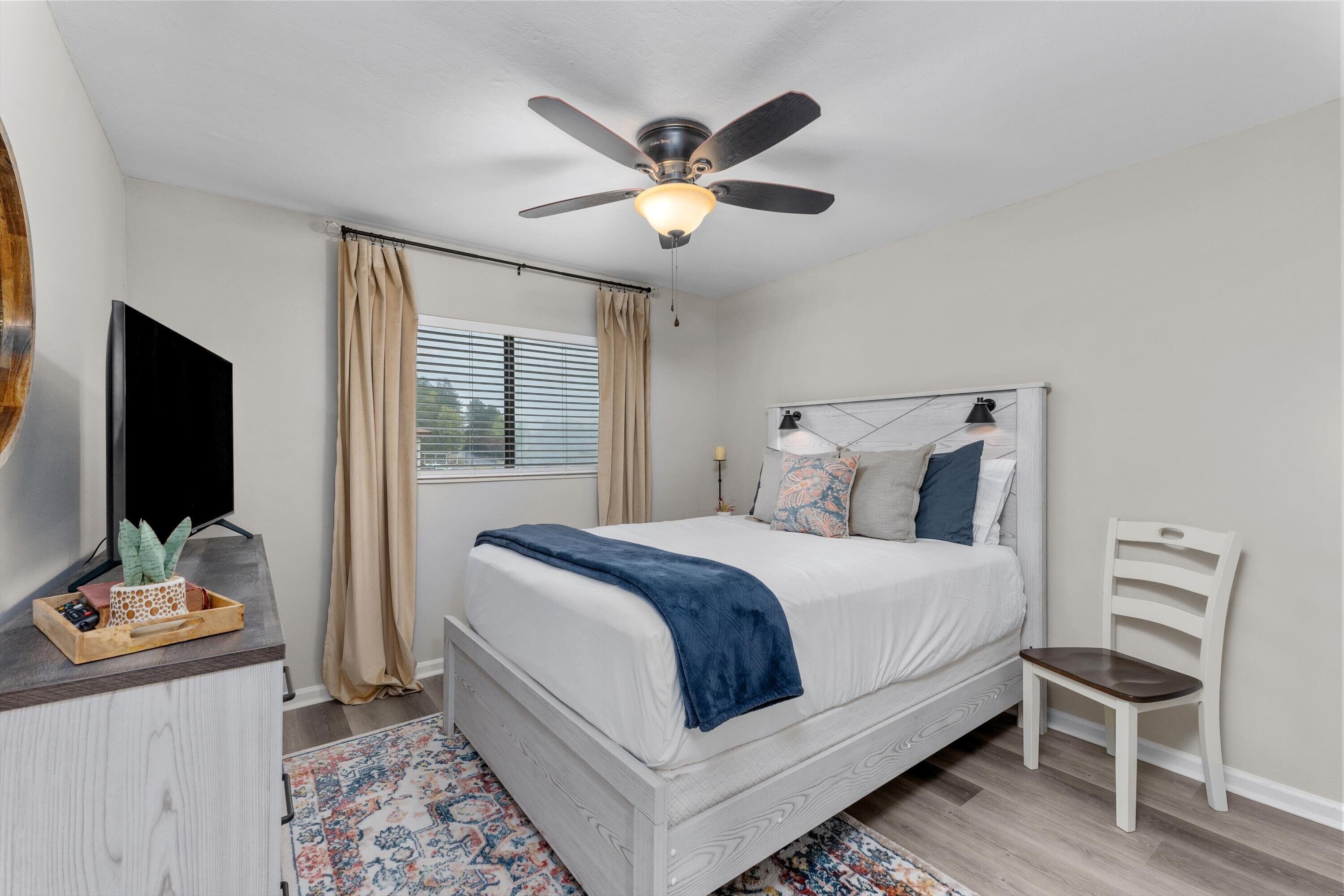 A bedroom with a bed, a ceiling fan, a TV, a desk, a chair, and a window with curtains.