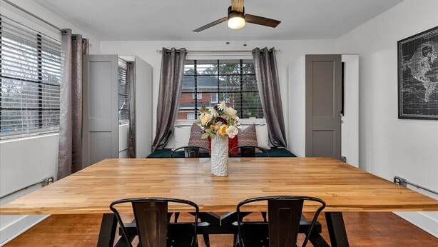Comedor con mesa de madera, sillas negras, ventilador de techo y un jarrón con flores.