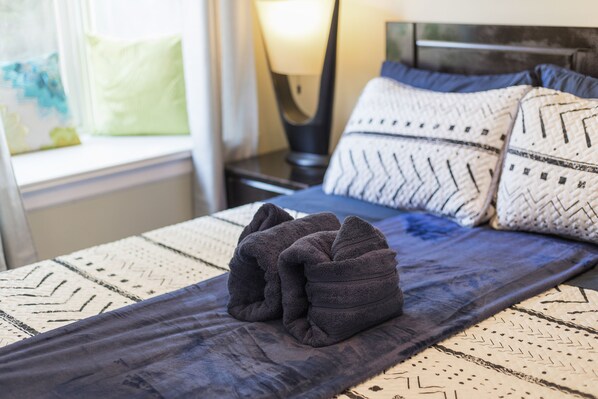 A neatly made bed with a dark blue blanket and two pillows, a bedside lamp, and a window with a view of the outside.