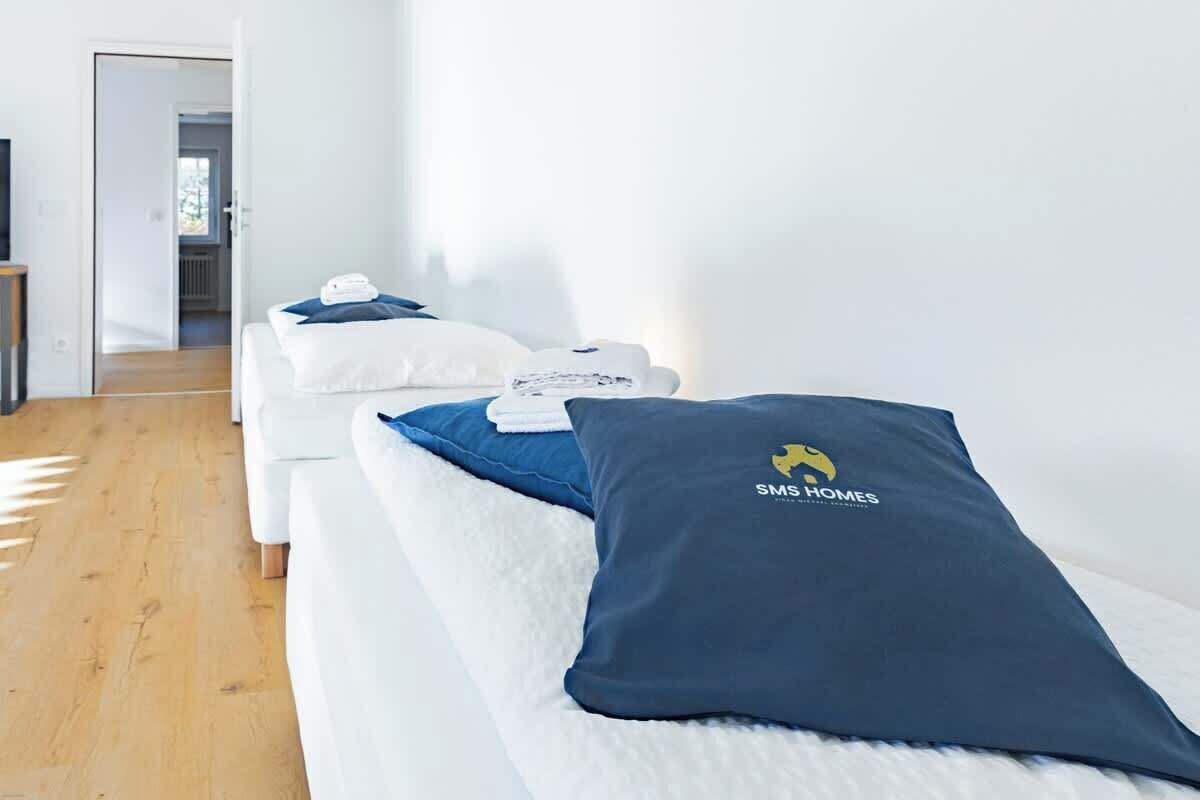 Una habitación de hotel con un sofá blanco, una almohada azul con el logotipo de SMS HOMES y una almohada blanca.