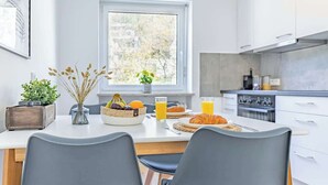 A modern kitchen with white cabinets, a dining table with two chairs, breakfast items, and a view of the outdoors through a window. - SMS-Homes I Timeout at the Alpsee-Lake (Immenstadt im Allgäu)