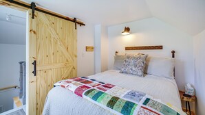 Un dormitorio con una puerta de madera, un edredón acolchado, una lámpara de pared y una mesita de noche con un control remoto.