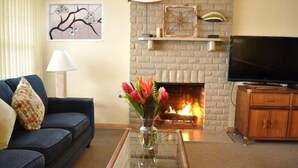Een woonkamer met een metselwerk open haard, een bank, een salontafel met een bloemenvaas, en een televisie op een houten kast.