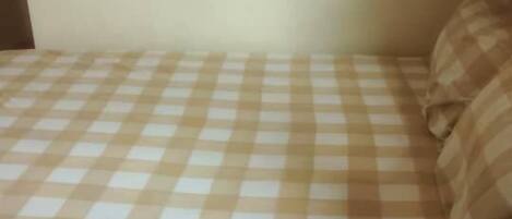 A small bed with a checkered bedspread, a bedside table with a lamp, and a wall-mounted shelf.