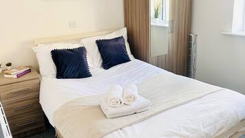 A neatly made bed with white linens and two dark blue pillows, a wooden nightstand with a few books, a large mirror, and a framed floral artwork on the wall.